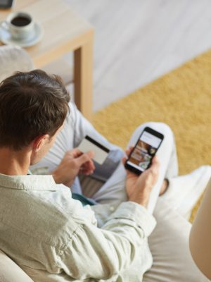 Rear view of young man sitting on sofa with mobile phone and shopping online using his credit card