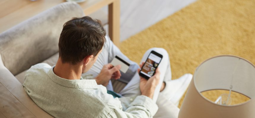 Rear view of young man sitting on sofa with mobile phone and shopping online using his credit card